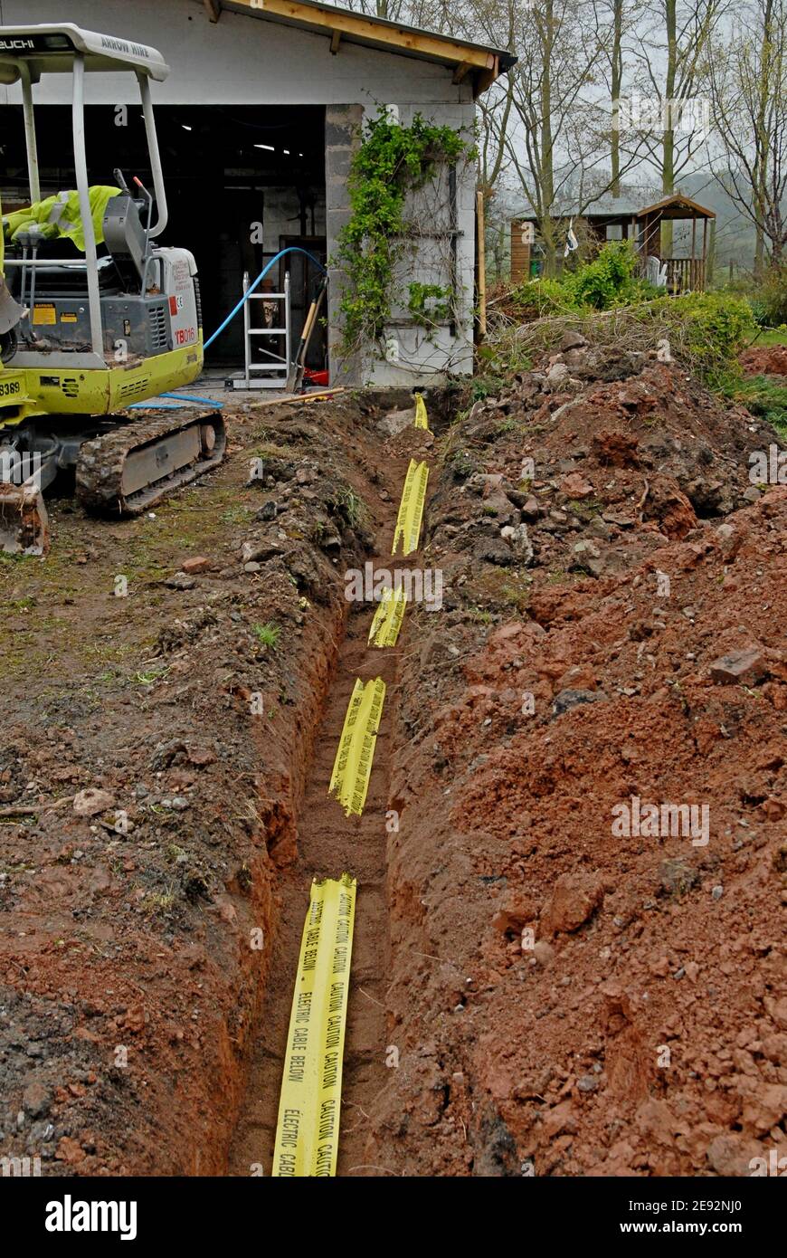 A mini digger stands next to a trench in a back garden with a newly laid electric cable and warning tape in the trench, Herefordshire, UK Stock Photo