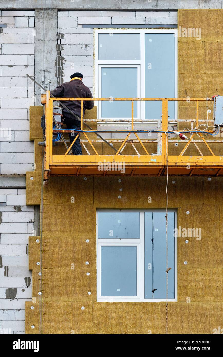 The builder stands on a suspended platform and insulates the facade of a house under construction. Stock Photo