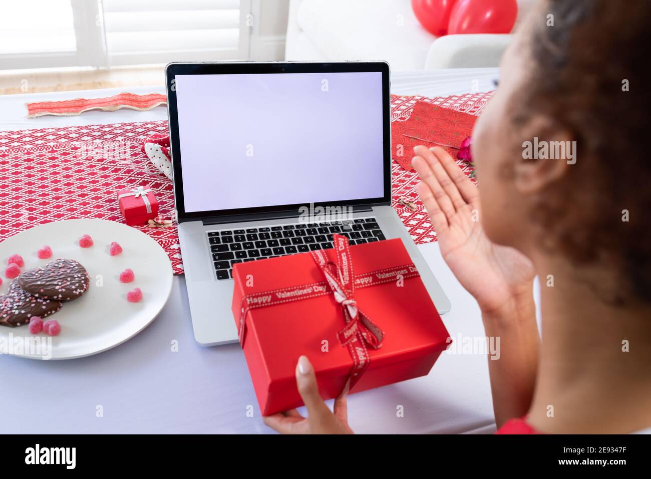 African american woman holding gift box blowing a kiss on videocall on laptop at home Stock Photo