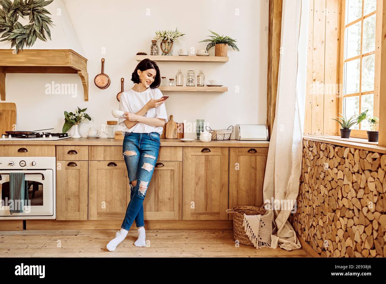 Lifestyle portrait of a happy young girl browsing the news on the phone over a cup of tea or coffee in the kitchen early in the morning Stock Photo