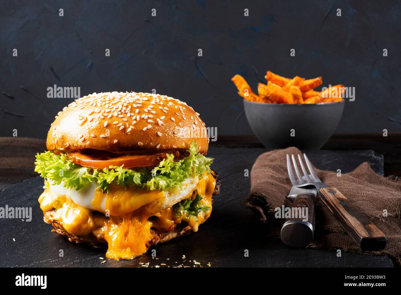 Juicy cheeseburger with egg arranged on a black skipper plate with cutlery and sweet potato fries, black background, text free space, Stock Photo