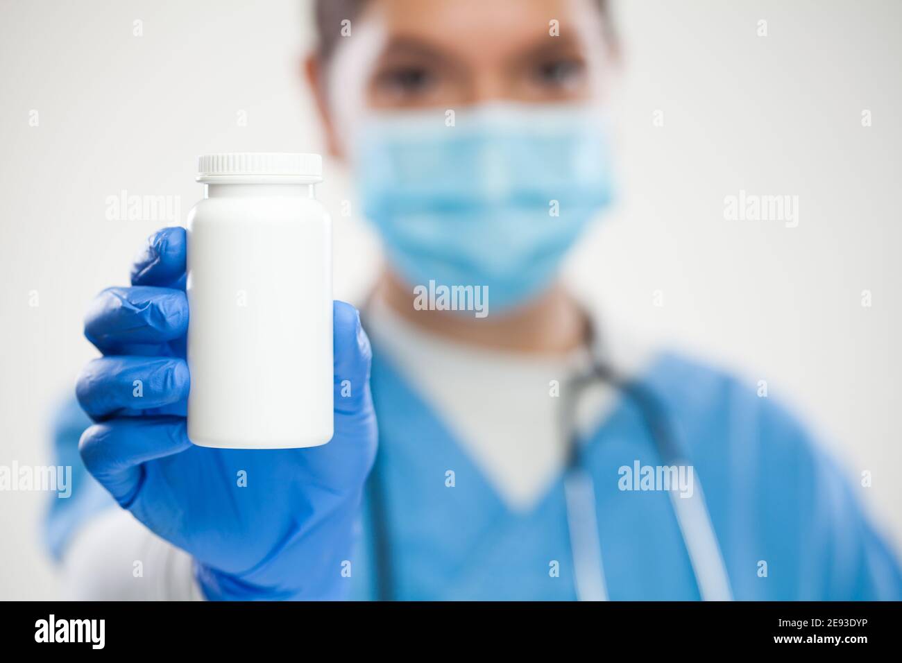 Female GP UK NHS general practitioner doctor,wearing scrubs,protective face mask and gloves,holding white blank bottle of pills,empty no label contain Stock Photo