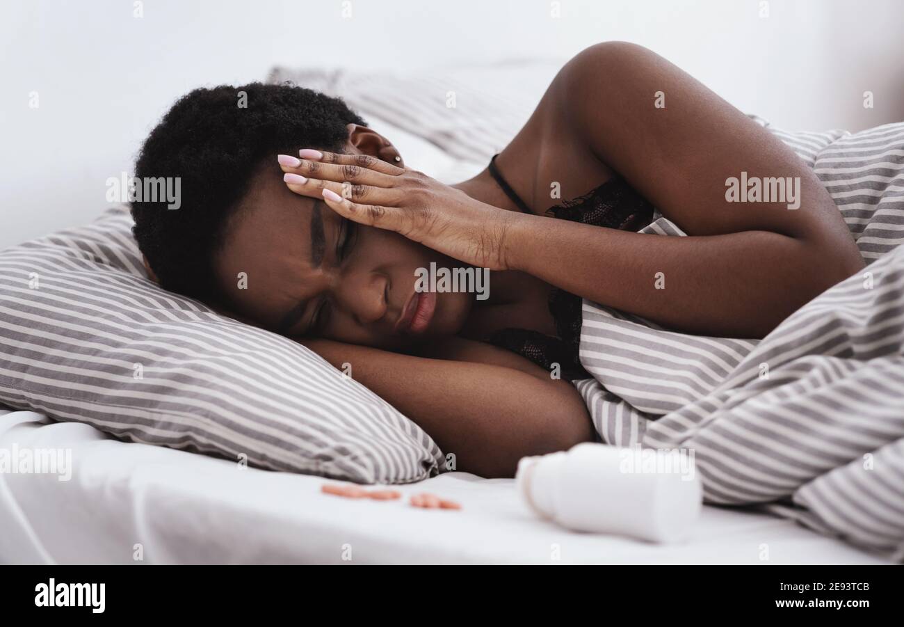 Ailing young african american lady with closed eyes presses hands to temples and suffers from headache lies on bed Stock Photo