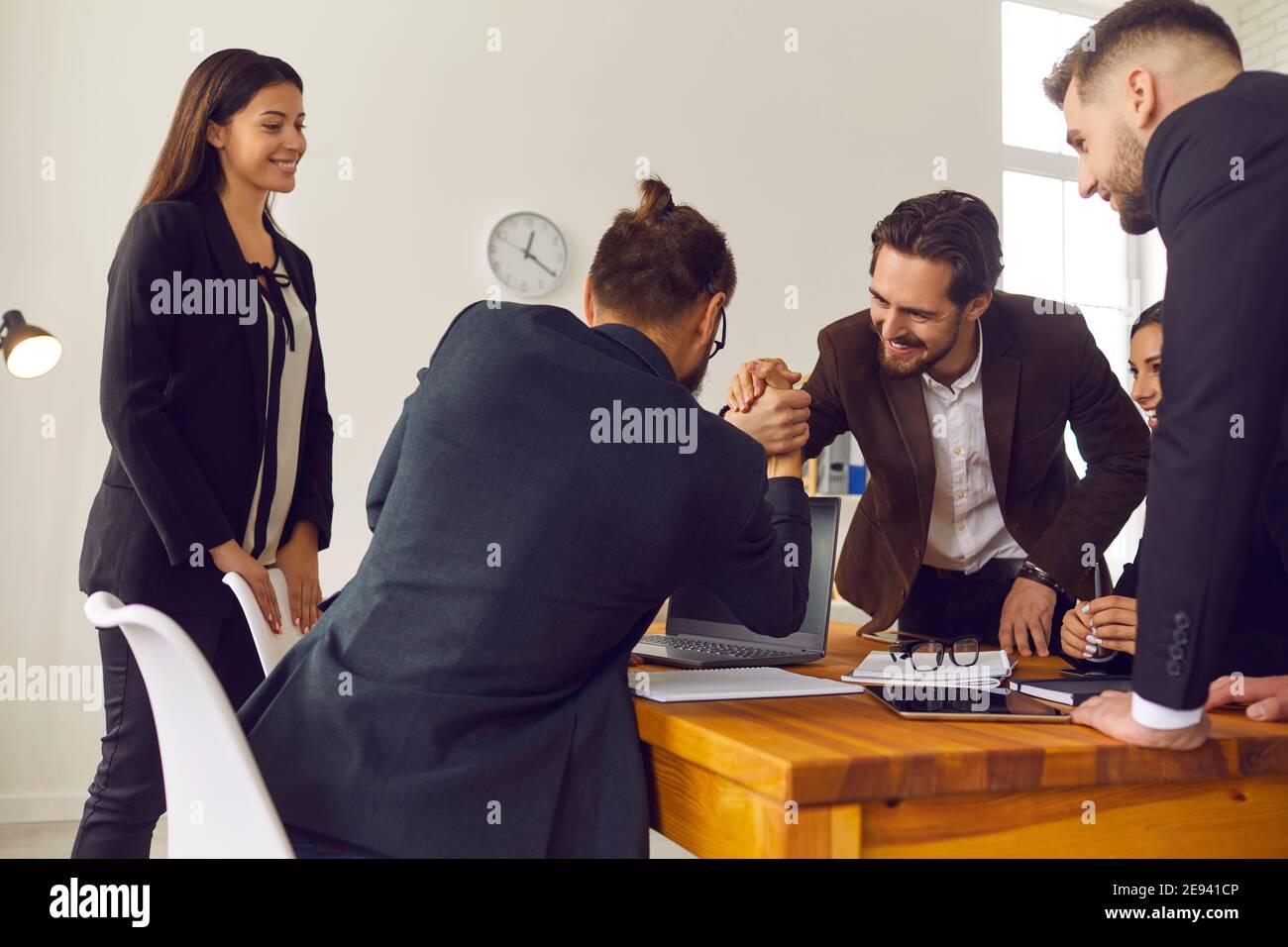 Two happy young businessmen shaking hands in office with smiling coworkers watching Stock Photo