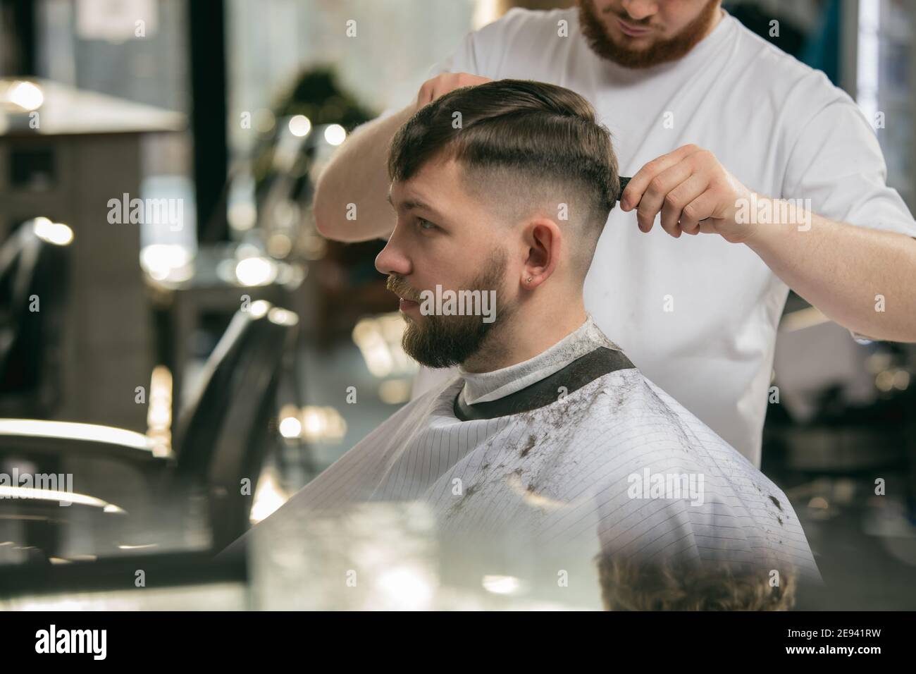 Close up master barber, stylist does the hairstyle to guy, young man. Professional occupation, male beauty concept. Cares of hair, mustache, beard of client. Soft colors and focus, vintage Stock Photo - Close up master barber, stylist does the hairstyle to guy, young man. Professional occupation, male beauty concept. Cares of hair, mustache, beard of client. Soft colors and focus, vintage Stock Photo -