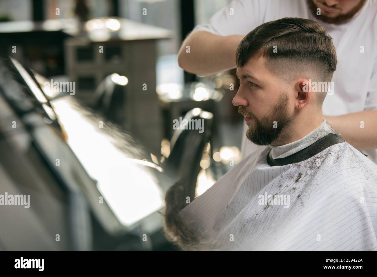 Close up master barber, stylist does the hairstyle to guy, young man.  Professional occupation, male beauty concept. Cares of hair, mustache,  beard of client. Soft colors and focus, vintage Stock Photo -