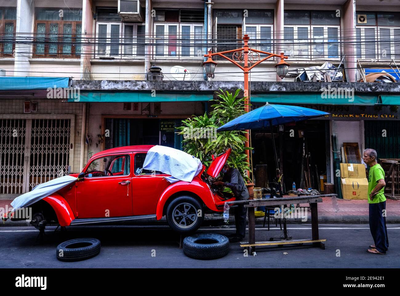 A man watches while a male car mechanic works on an old VW Beetle in a street in the Chinatown area of Bangkok, Thailand Stock Photo