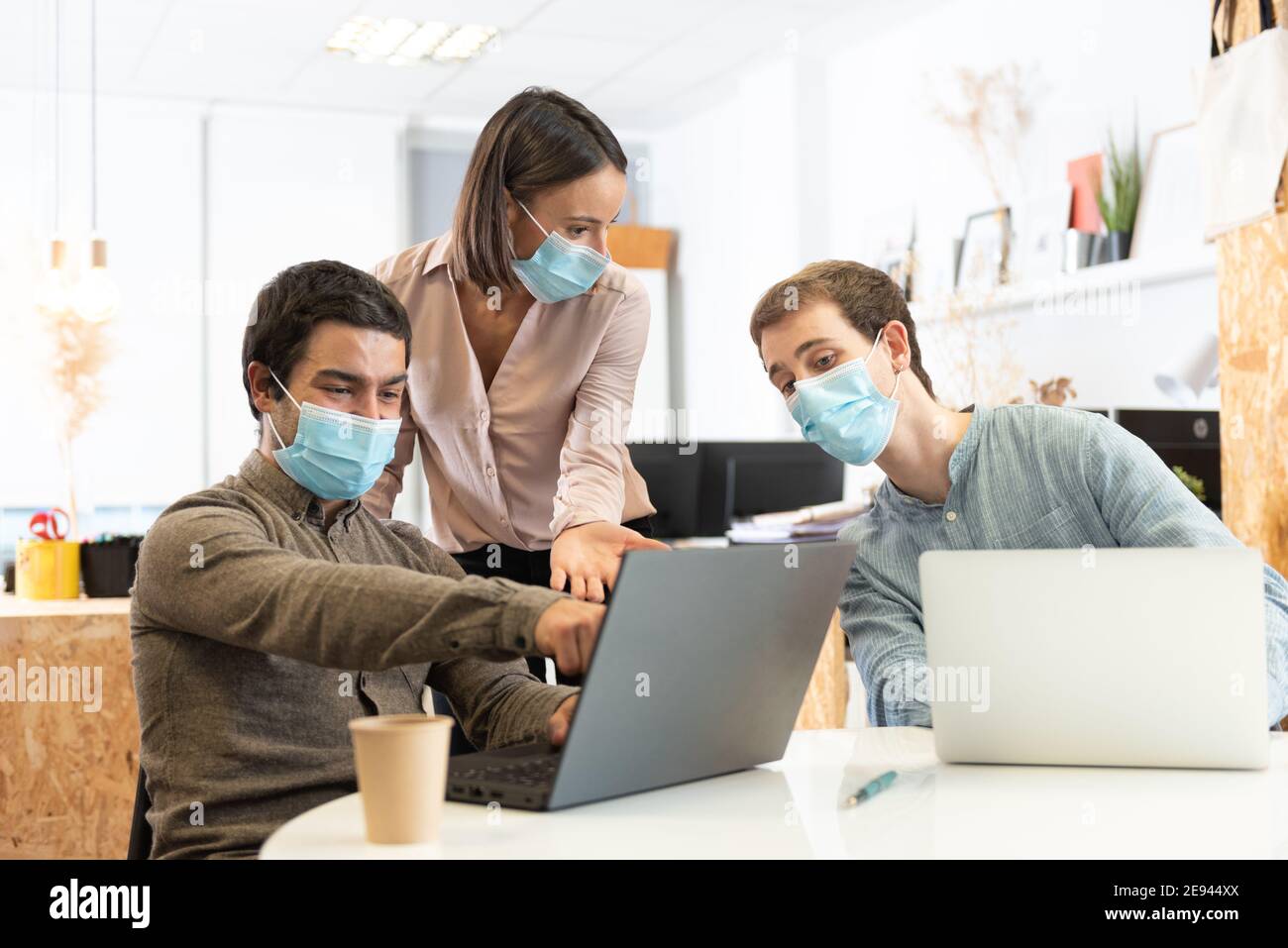 Three coworkers discussing and looking at a laptop while wearing face masks. Working in the office during Coronavirus pandemic concept. Stock Photo