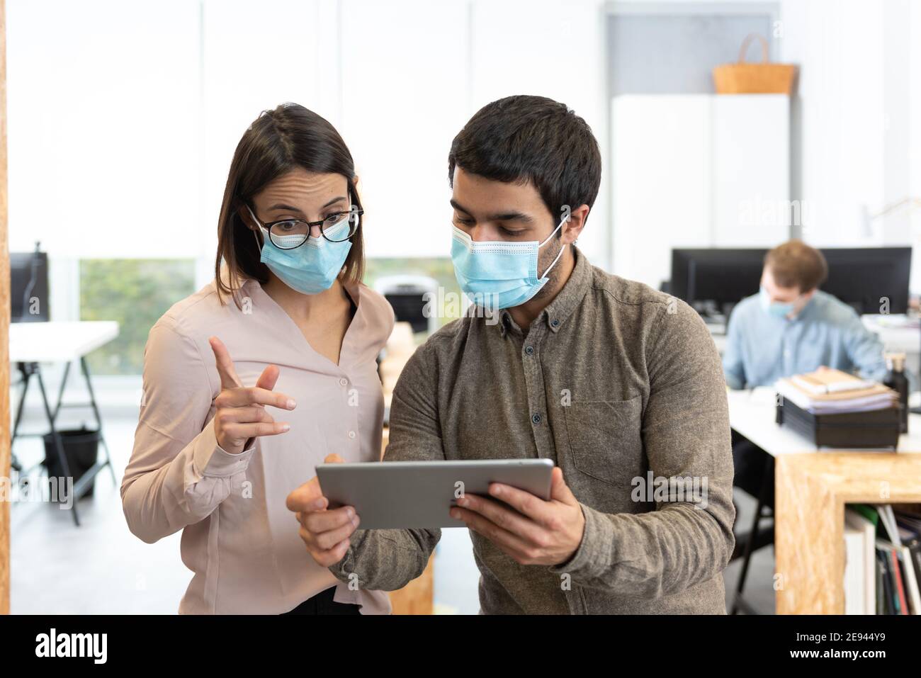 Hispanic man and woman wearing protective face masks chatting while looking a tablet. Working in the office during Coronavirus pandemic concept. Stock Photo