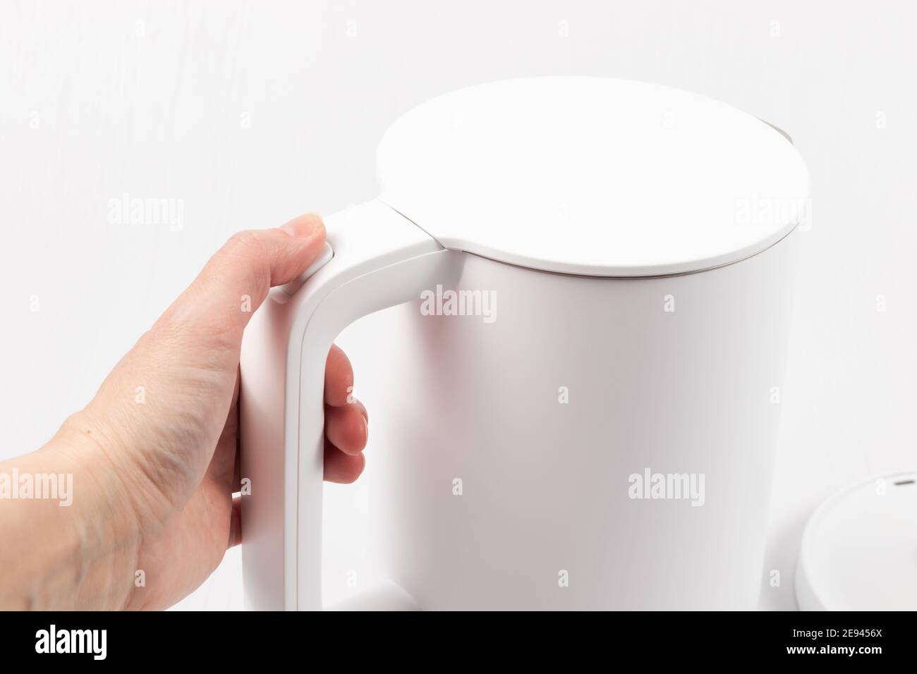 a female hand holds a white electric kettle by the handle on a white wooden table background Stock Photo