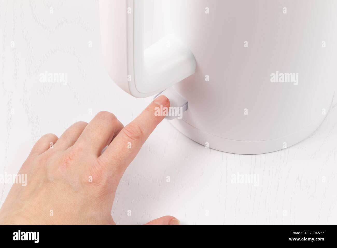 A Female Hand Turns On An Electric White Kettle To Boil Water On The a-female-hand-turns-on-an-electric-white-kettle-to-boil-water-on-the