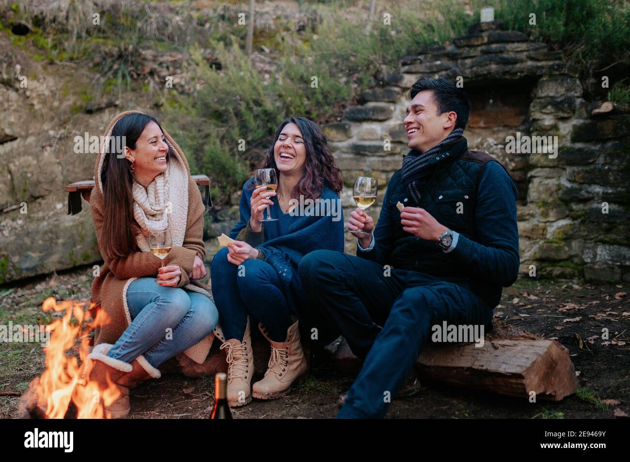 Group of happy friends sitting near a bonfire drinking glass of ...