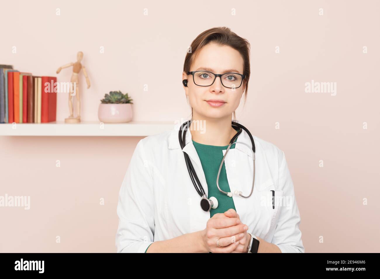 Portrait of confident young female doctor wearing white coat with stethoscope and earpiece standing in front of a camera at her home office performing Stock Photo
