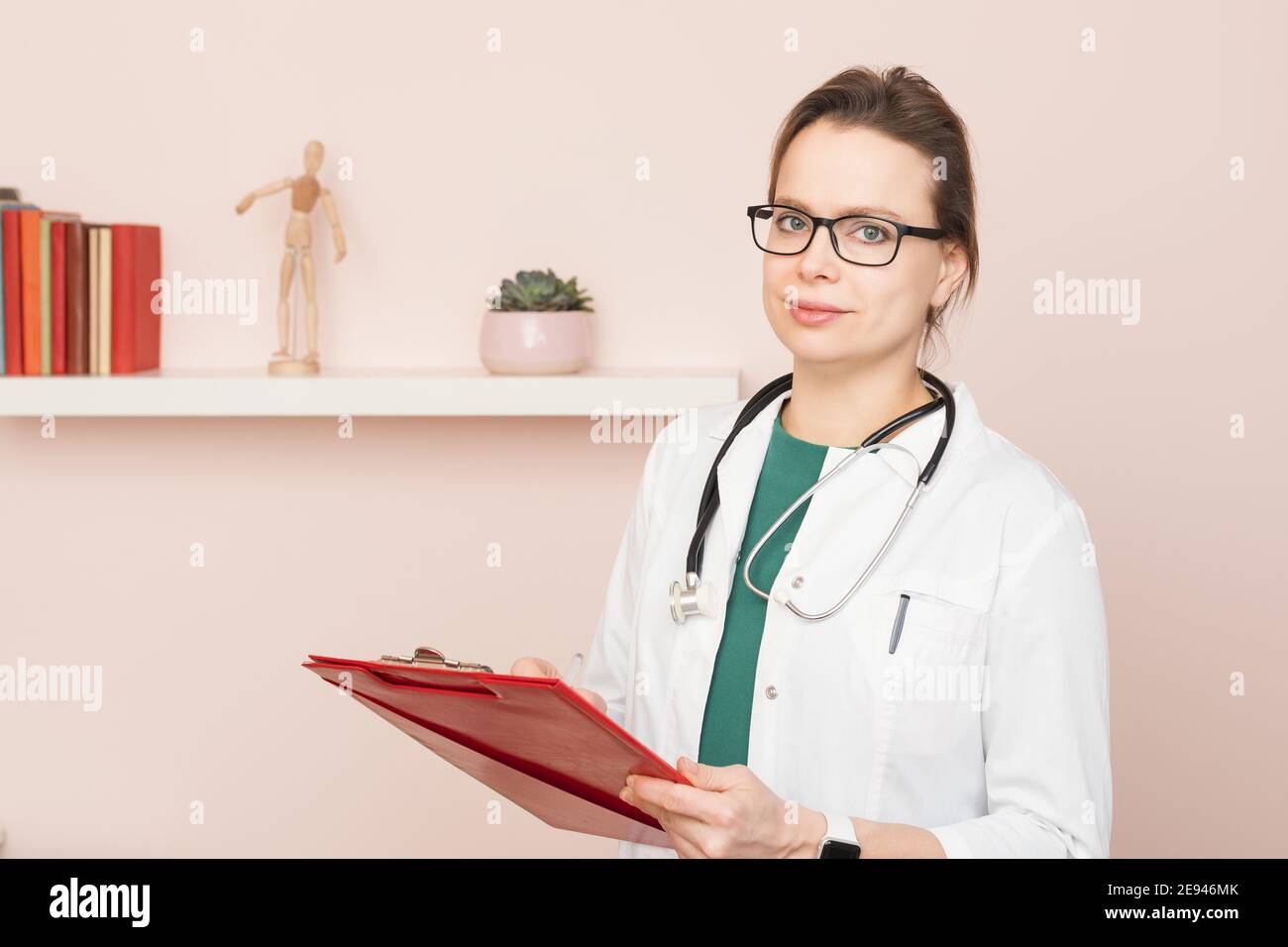 Portrait of confident young female doctor wearing white coat with stethoscope standing in front of a camera at her home office filling medical form pe Stock Photo