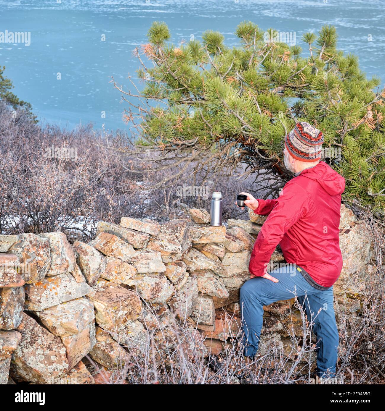 senior hiker is taking a tea break at old stone fence overlooking frozen mountain lake  - Horsetooth Reservoir at foothills of Rocky Mountains - a pop Stock Photo