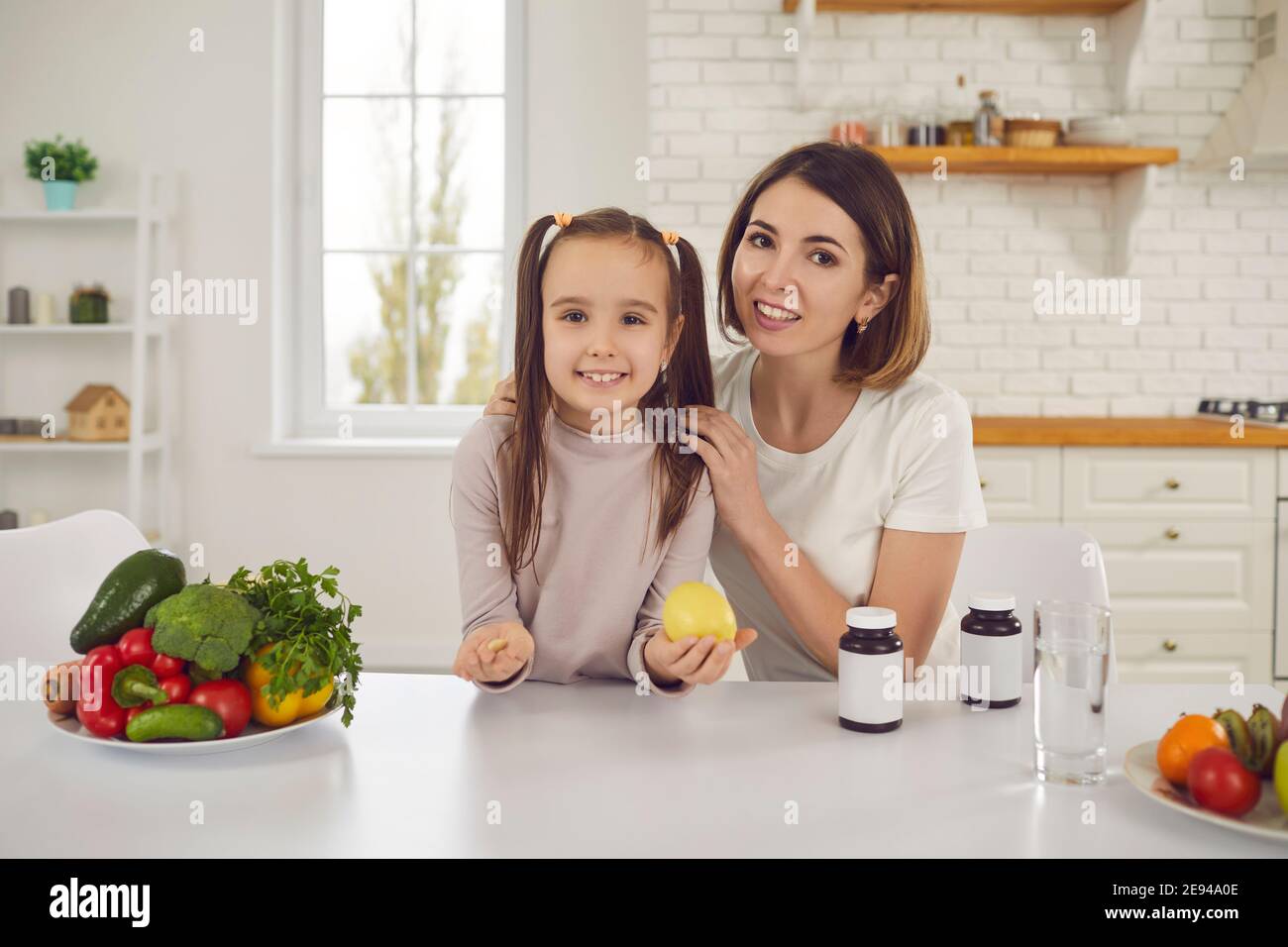 Happy mom and daughter at kitchen table with fruit, veggies and food supplements Stock Photo