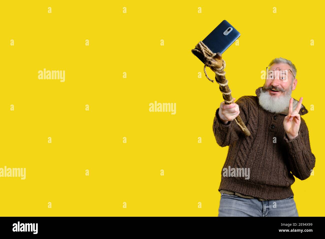 Studio portrait of mature bearded man taking selfie with wooden stick, funny selfie making concept, yellow background Stock Photo
