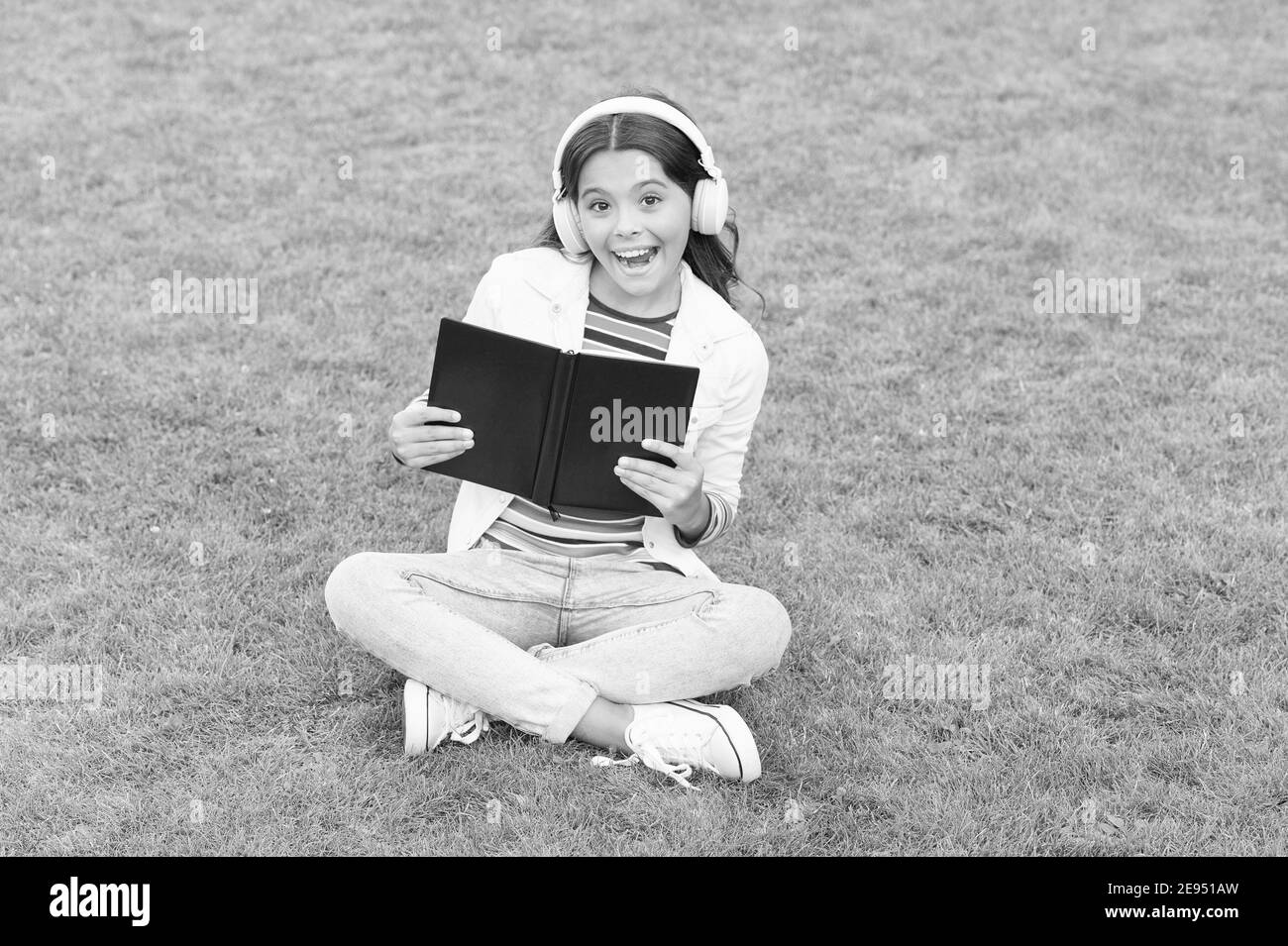 Work hard. small girl reading book. literature for kids. write childhood memories. smart kid in headset. back to school. kid study online in park. listen music on green grass with book. Stock Photo
