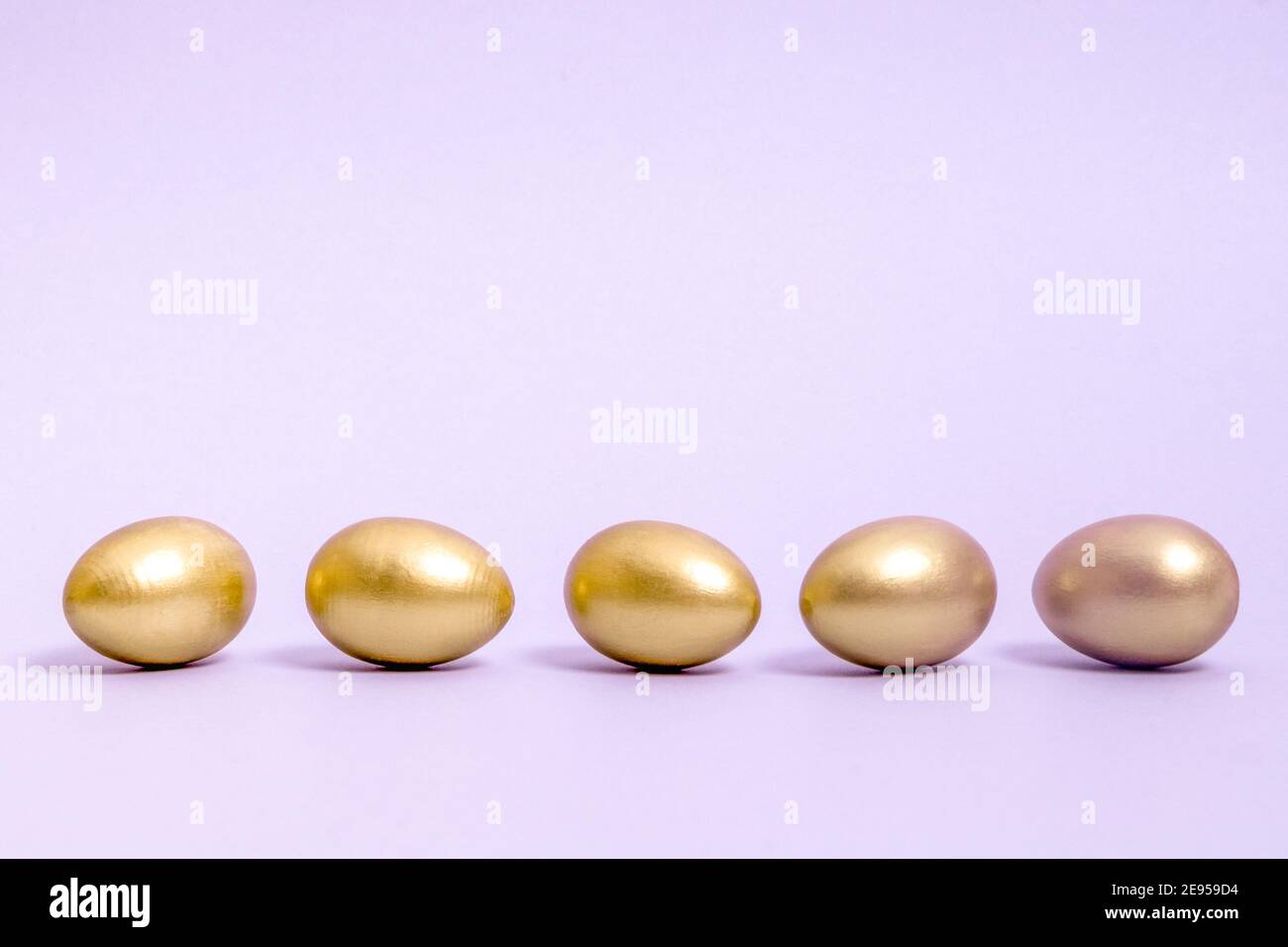 Five Easter eggs decorated with gold paint lie in a row against a lavender background. Concept for Easter, spring. Selective focus. Copy space. Stock Photo