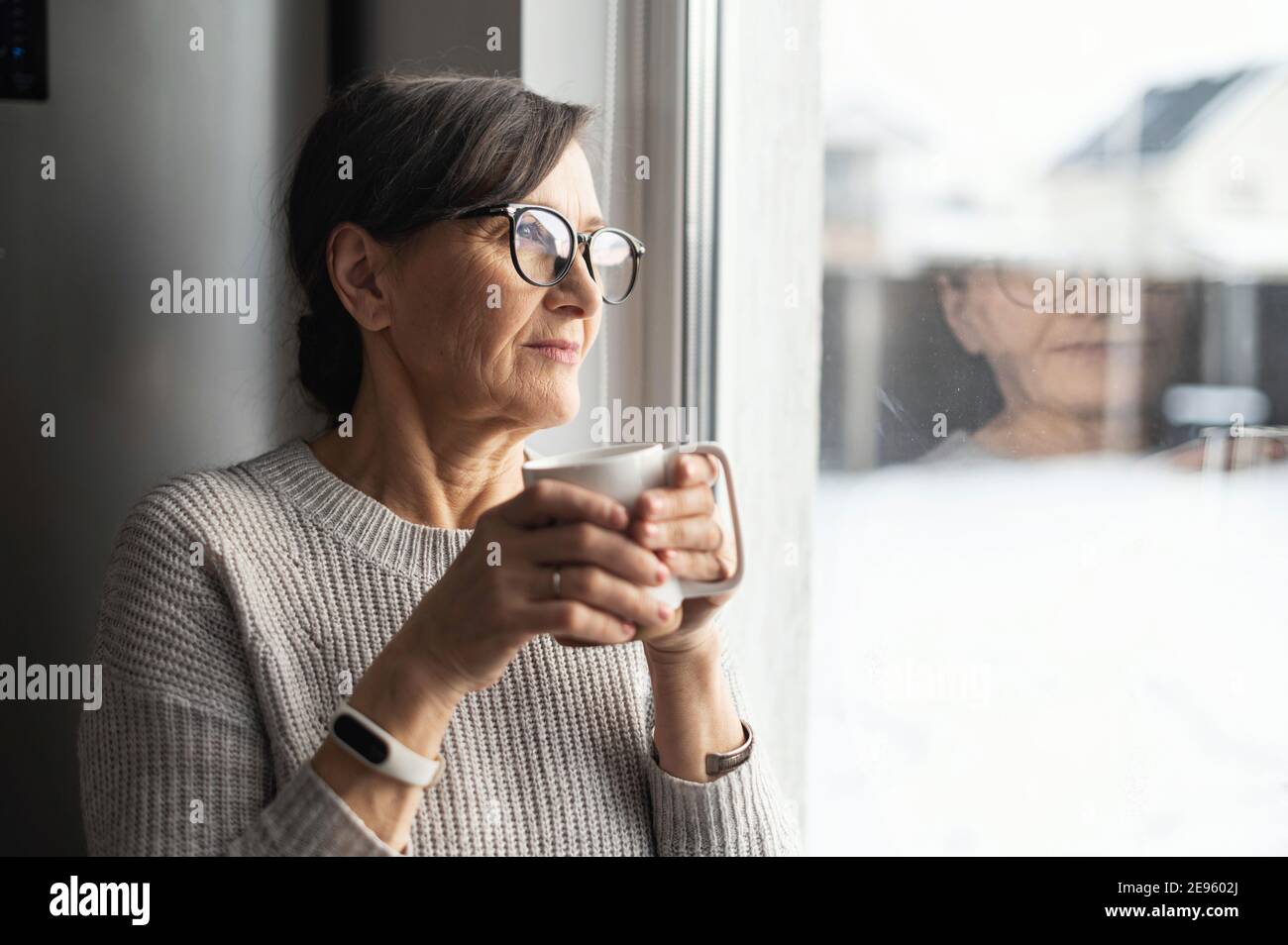 Close-up portrait of senior older woman wearing glasses enjoys morning coffee in the kitchen at home. A modern retirement lady daydreaming with a mug Stock Photo