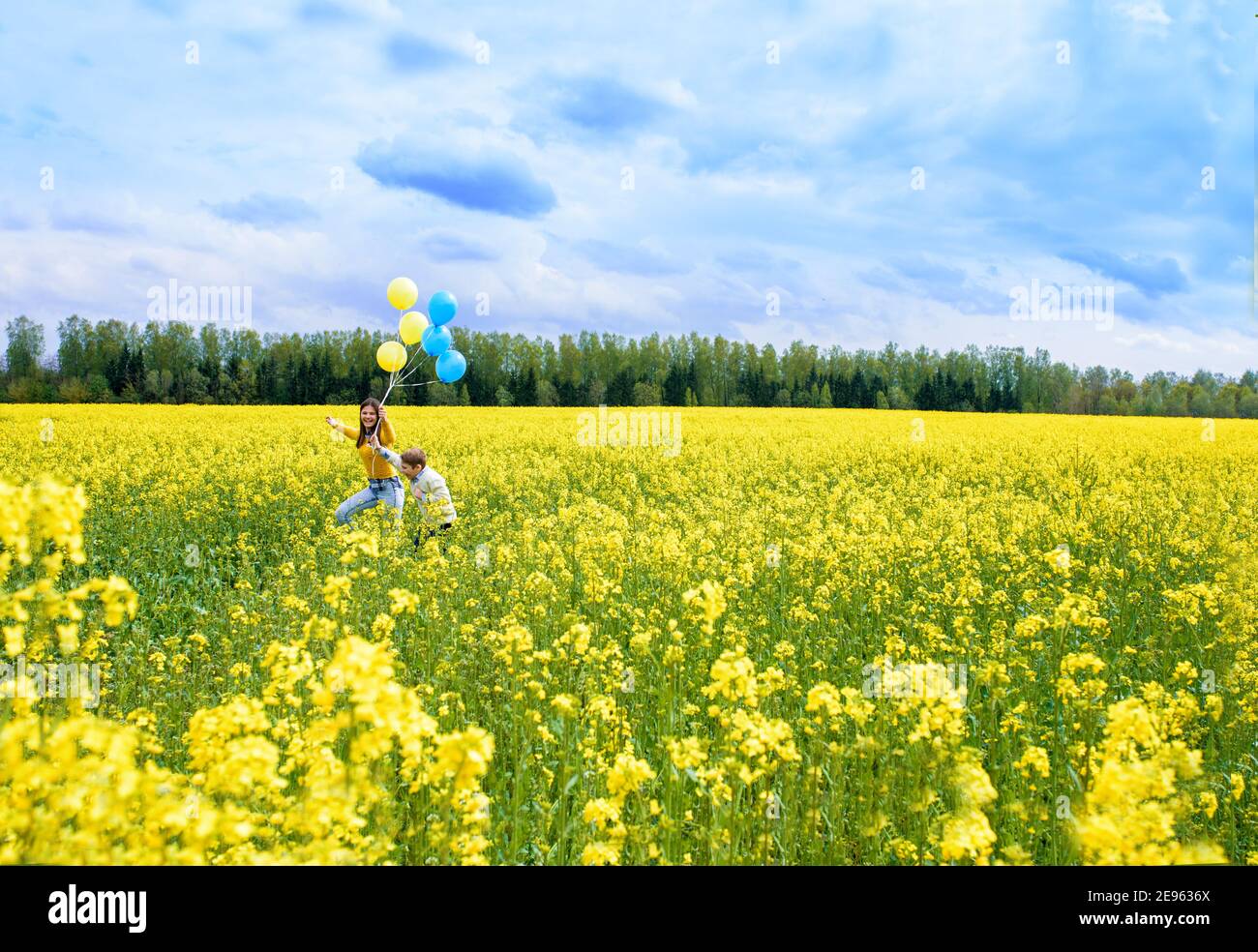 happy two children run on a yellow field, blooming rapeseed. blue sky and clouds. Blue and yellow balloons, concept of freedom, summer. Holiday and Stock Photo