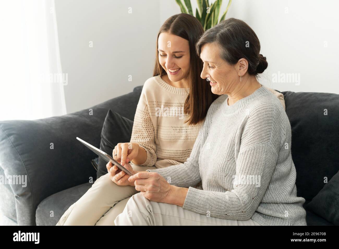 Side view an adult daughter and a senior mother spend time with a tablet indoor, a young woman showing to mature mom smth in a network, helps to Stock Photo