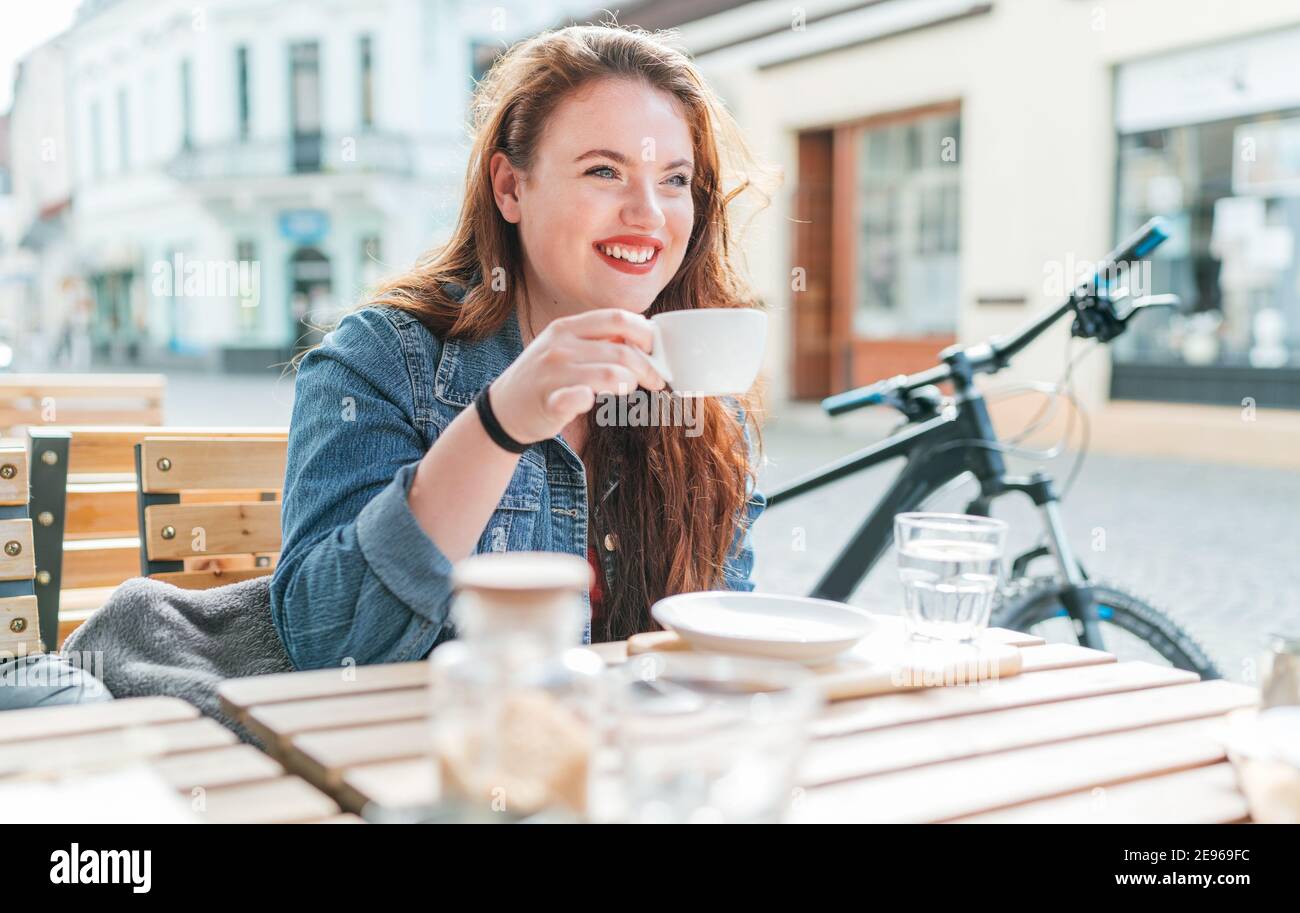Portrait of sad red curled long hair caucasian teen girl sitting on a cozy cafe outdoor terrace on the street and drinking coffee. Young woman taking Stock Photo