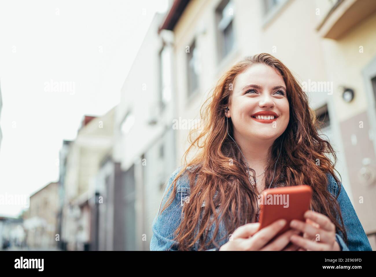 Smiling red curly long hair caucasian teen girl walking on the street and browsing the internet using the modern smartphone. Modern people with techno Stock Photo