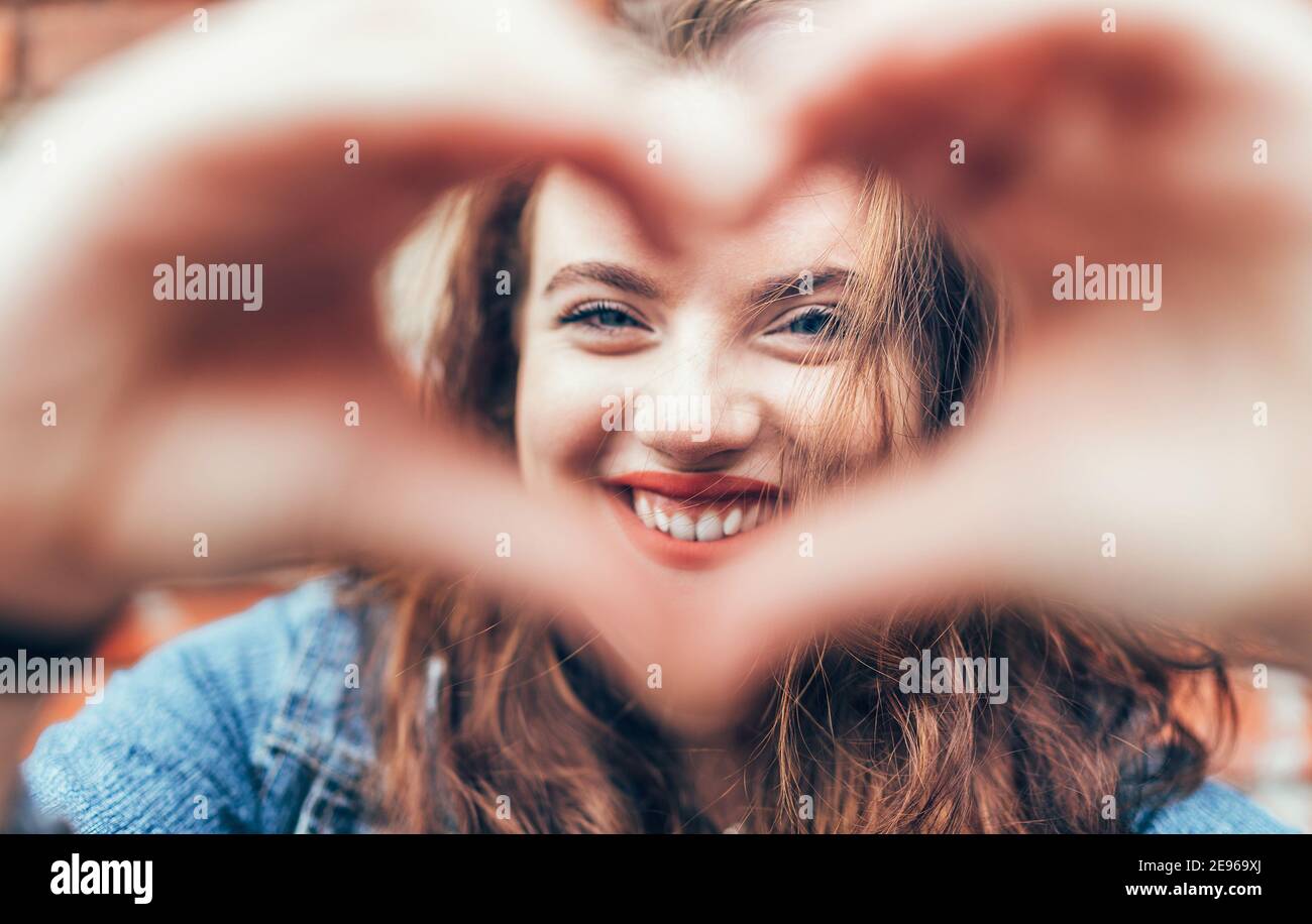 Portrait of sincerely smiling red curled long hair caucasian teen girl with applied red lipstick lips with blue eyes making a heart shape with fingers Stock Photo