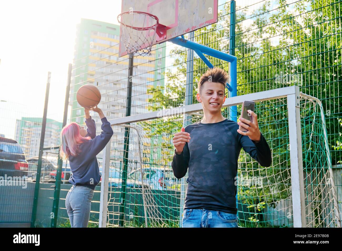 Guy teenager recording video stream using smartphone, on sports street court, girl playing basketball Stock Photo