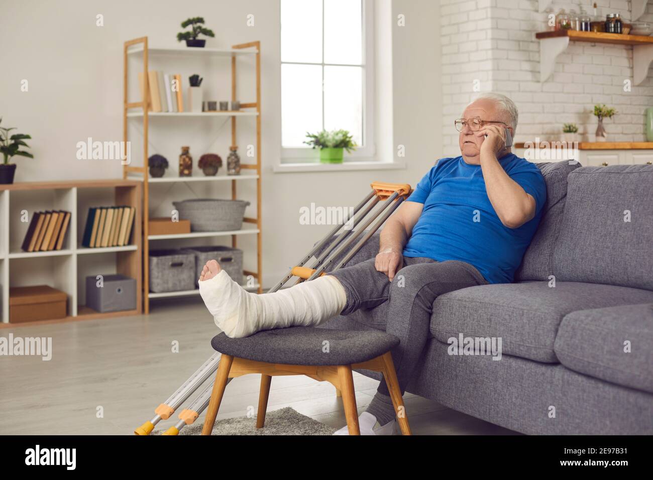 Senior man with leg fracture sitting on couch at home and talking on mobile phone Stock Photo
