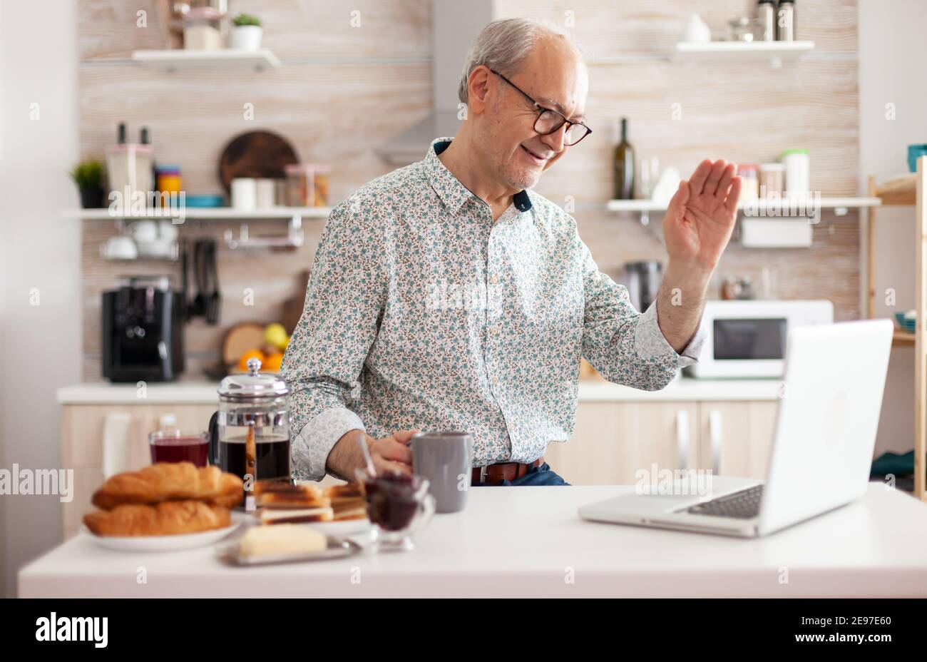 Daily life of senior man in kitchen during breakfast using laptop holding a cup of coffee, talking online with nephews waving at webcam. Retired person working from home on virtual meeting, conference Stock Photo