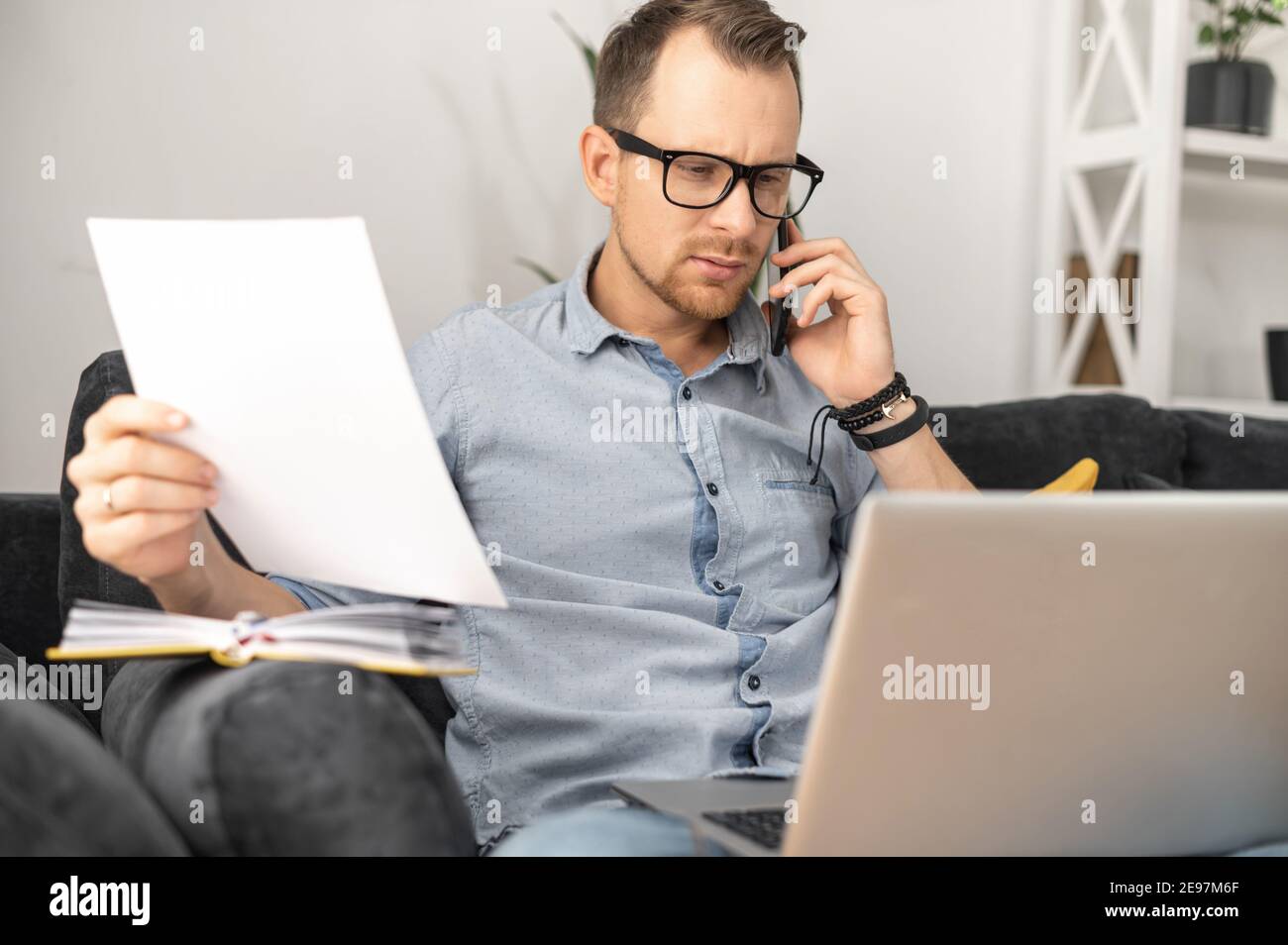 An intelligent busy businessman wearing eyeglasses working on laptop from home, talking on smartphone and holding documents, doing paperwork, checking Stock Photo