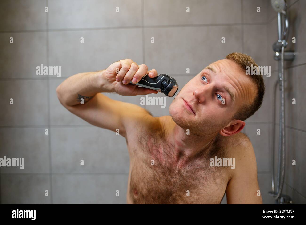 A man shaves his face with an electric razor in front of a mirror. Skin irritation. Bath procedure Stock Photo