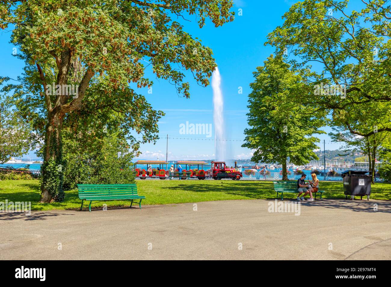 Geneva, Switzerland - Aug 15, 2020: Red tourist train with carriages and locomotive along the promenade du Lac in Jardin Anglais with Jet d'eau Stock Photo