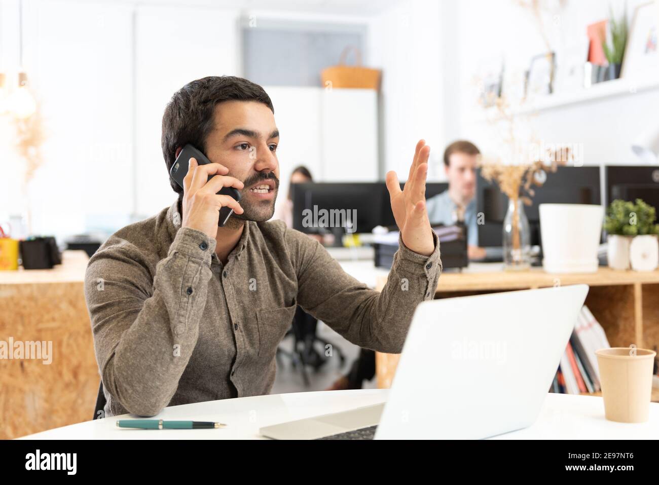 Angry hispanic man with mustache discussing on the phone in front of his laptop in the office. Stock Photo