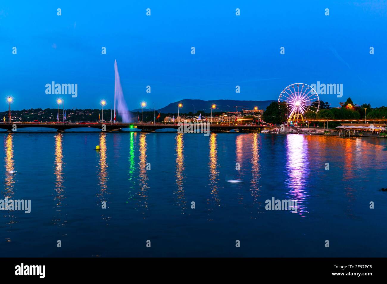 The Swiss Ferris Wheel and Pont du Mont-Blanc reflecting on Rhone river overlooking Lake Leman, French Swiss. Night view of Jet d'eau fountain and Stock Photo