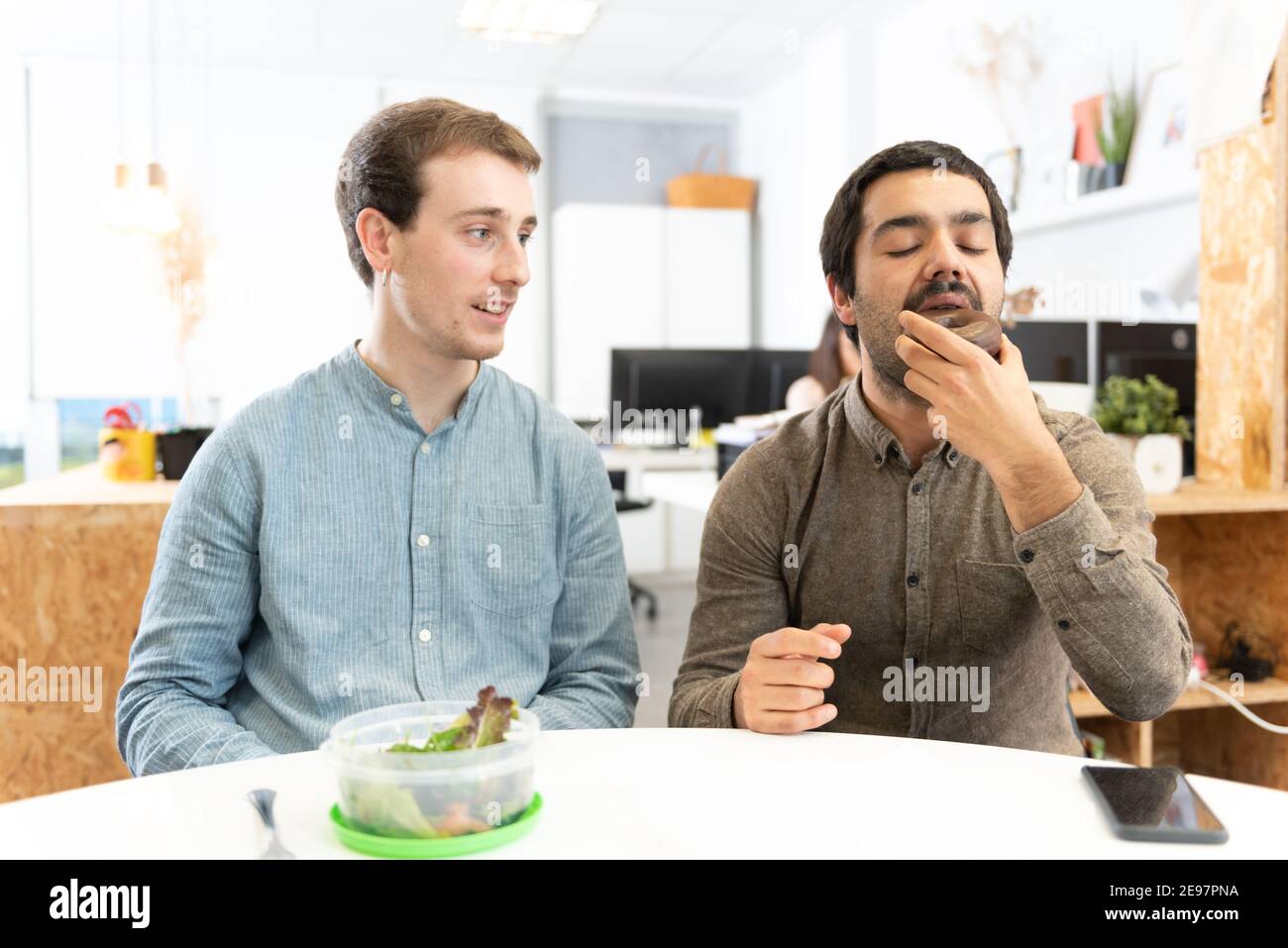 A man with a salad looking with envy to his coworker eating a sweet food in the office. Stock Photo