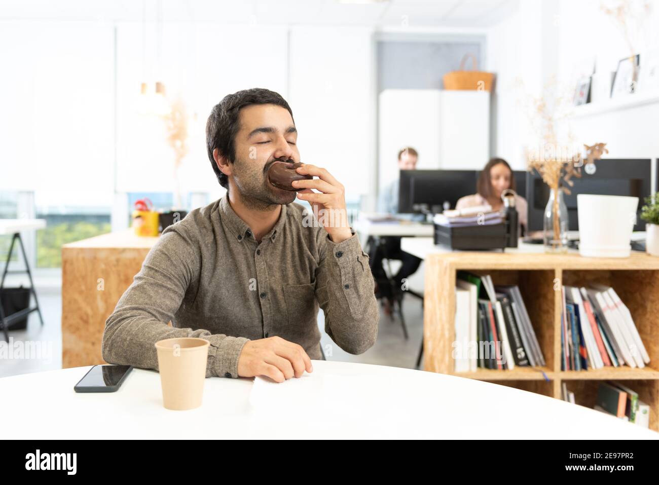Hispanic man with mustache eating and enjoying a chocolate sweet bun. Unhealthy habits in the office concept. Stock Photo