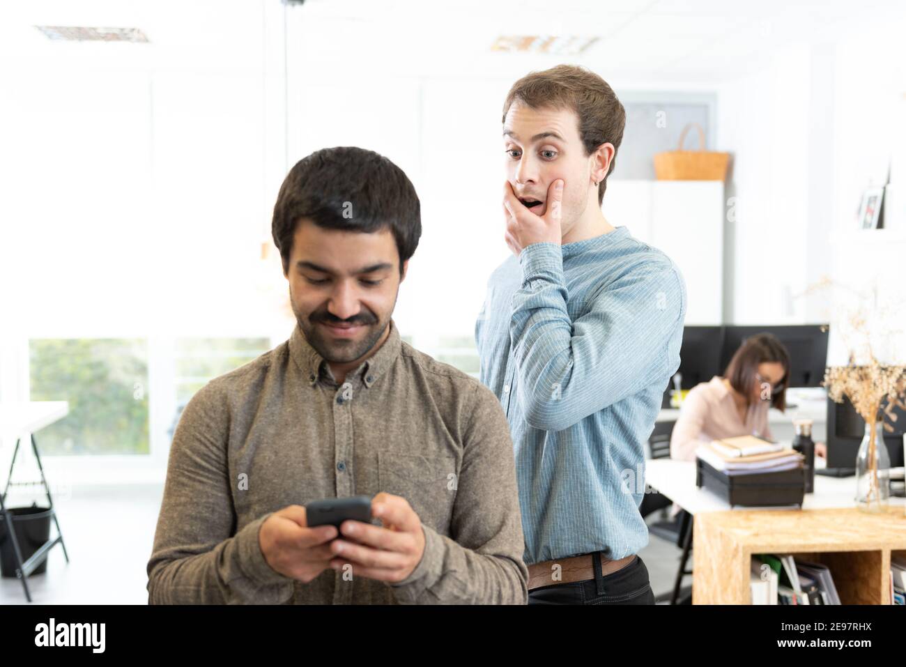 Surprised young man spying a coworker while he is looking at his cell phone at the office. Stock Photo