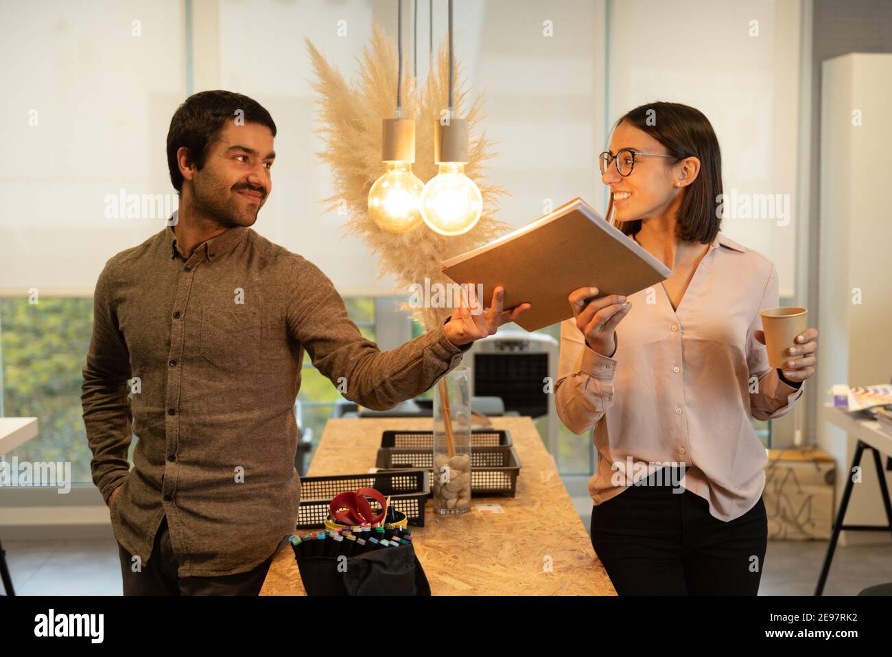 Happy hispanic man giving a folder to his female smiling coworker while looking each other. Good working environment concept. Stock Photo