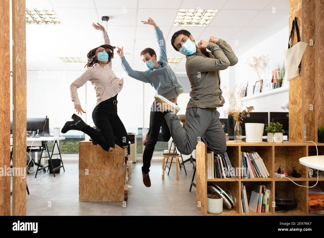 Happy coworkers wearing protective masks jumping and celebrating at the workplace. Working in the office during Coronavirus pandemic concept. Stock Photo