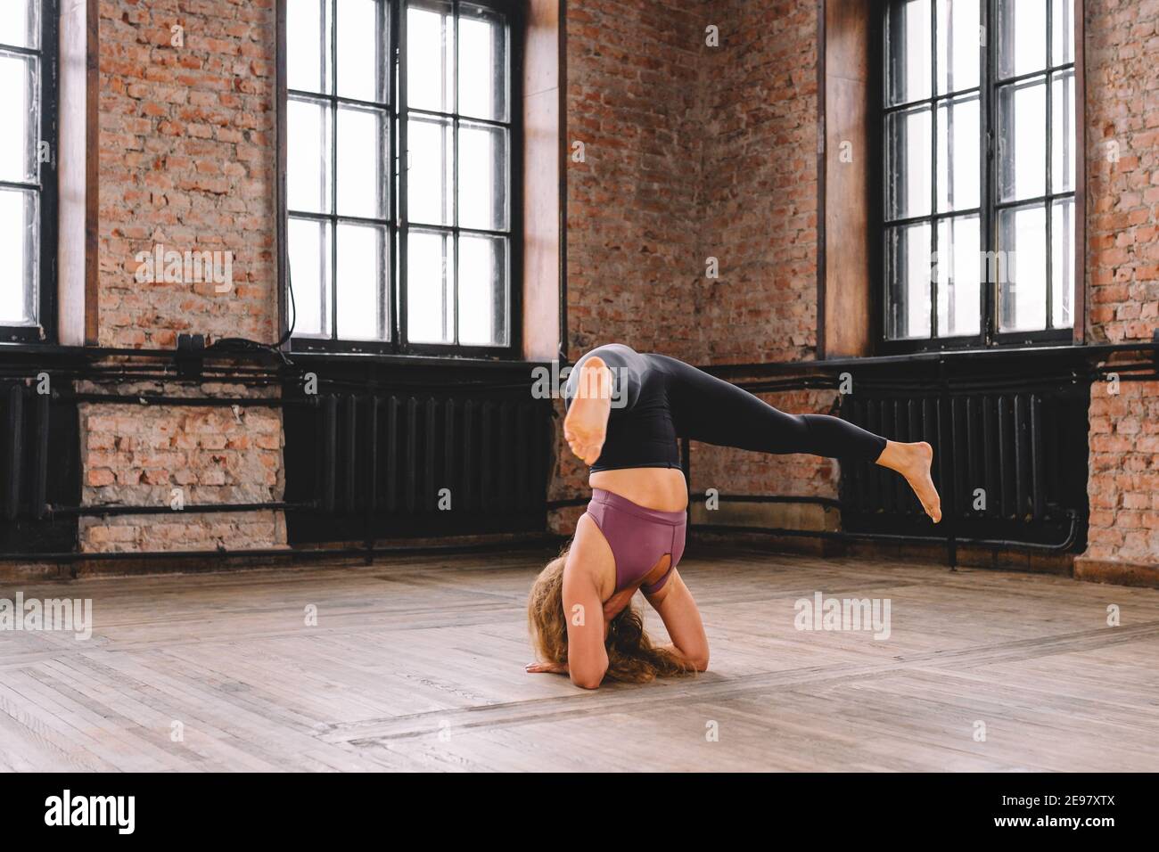 Young curly female do complex of stretching yoga exercises on floor of loft hall and smile. Positive, meditation, concentrate. Stock Photo