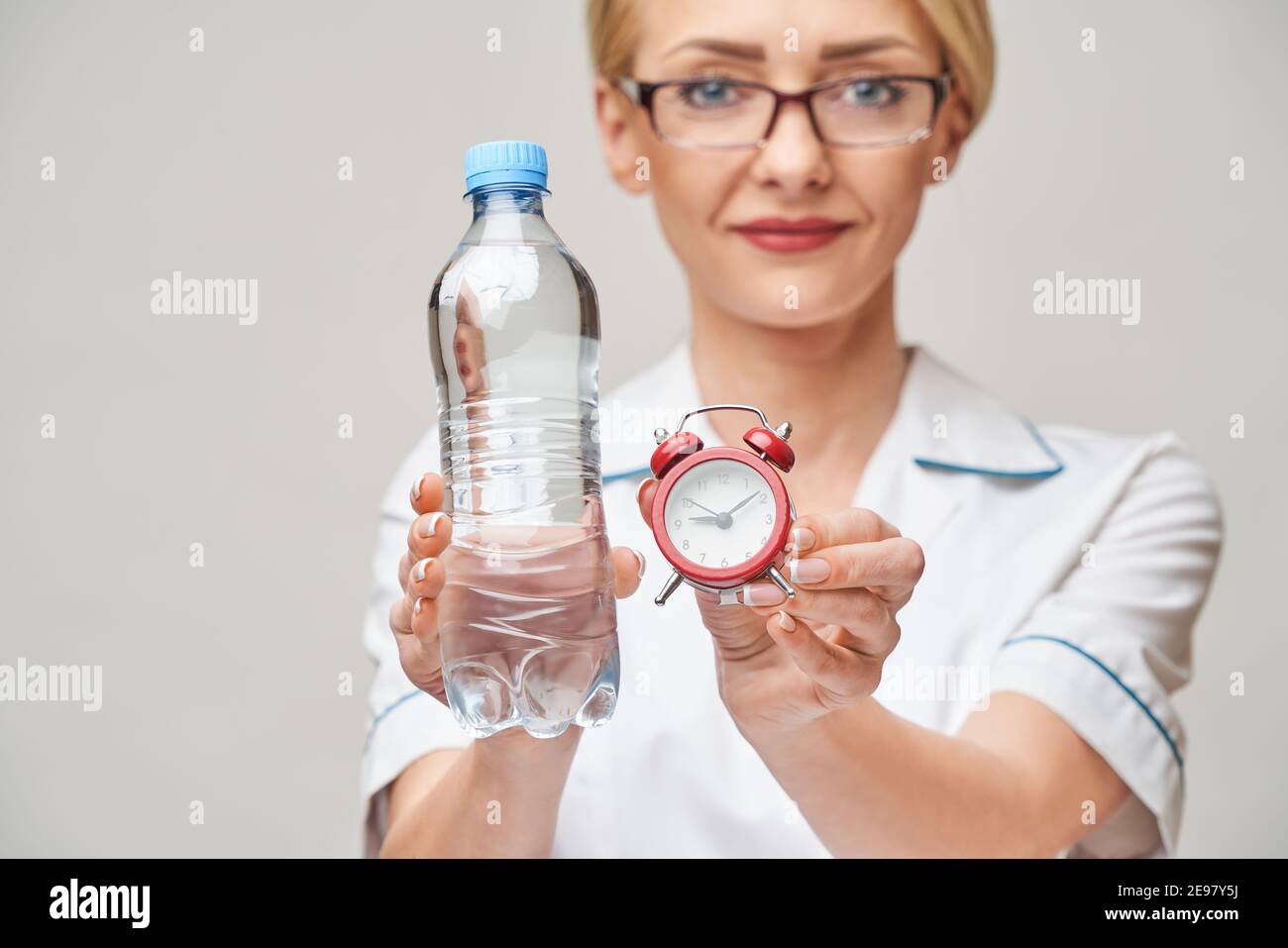 smiley nurse or doctor pointing holding small red alarm clock and bottle of water satanding over