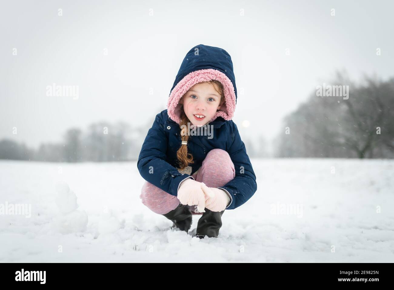 Beautiful young girl smiling happy playing outside in falling snow looking at camera wrapped up warm with blue coat hood up gloves and boots snowfall Stock Photo