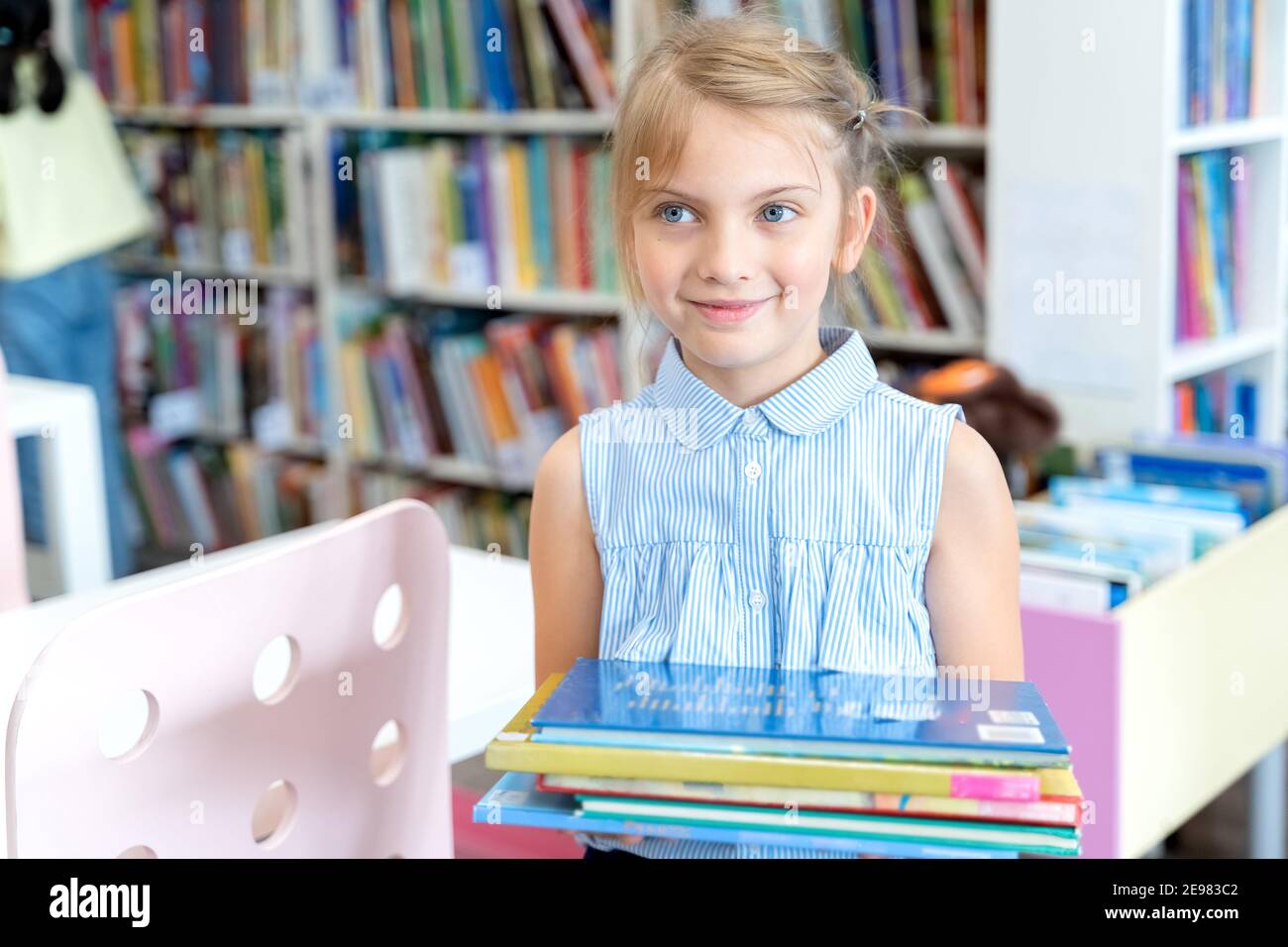 Little girl holds stack of books with fairy tales in children's library. Special reading kids room. Shelves with bright interesting, educational books Stock Photo