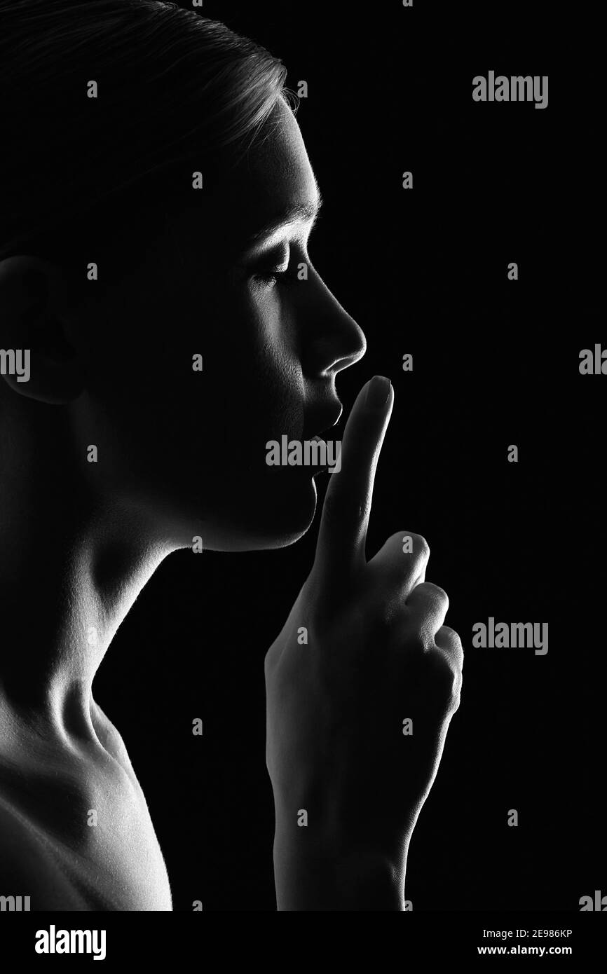 serious young woman with finger on her lips show silence gesture on black background, profile view, monochrome Stock Photo