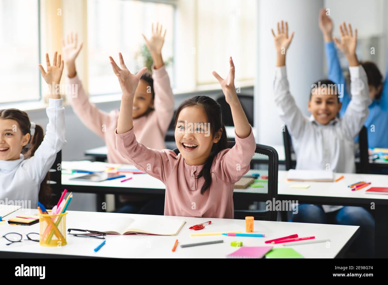 Happy diverse small schoolchildren raising hands at classroom Stock Photo