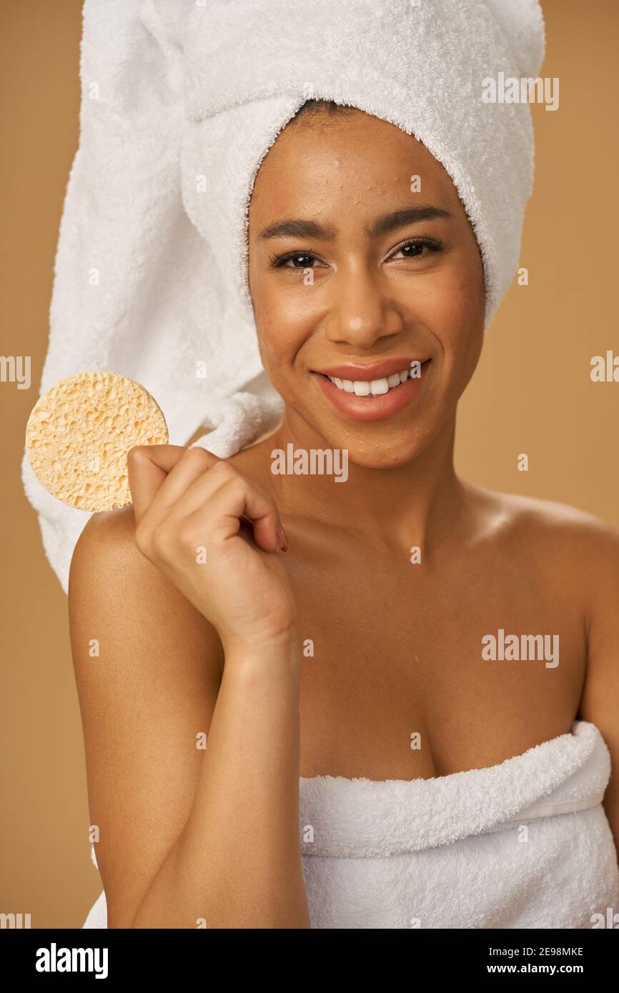 Funny mixed race young woman smiling at camera, holding cleansing face sponge, posing isolated over beige background. Bathroom routine. Youth and skin care concept. Vertical shot Stock Photo