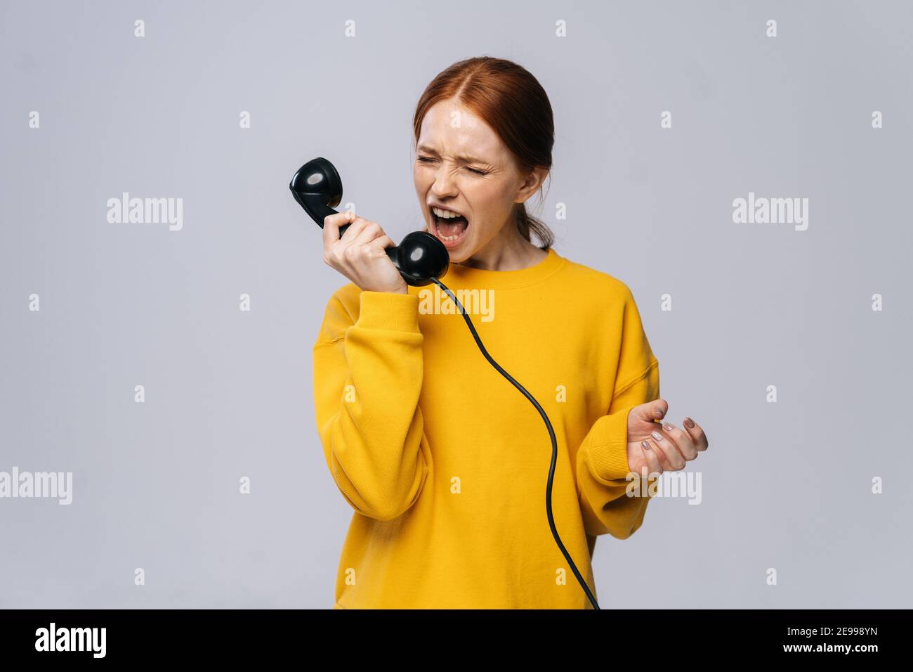 Crazy angry young woman in stylish yellow sweater talking on retro phone and screaming in handset. Stock Photo